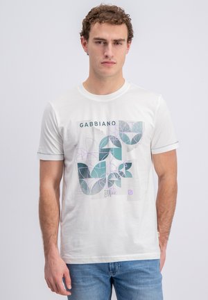 Man draagt wit T-shirt met korte mouwen met abstract blauw-purper geometrisch plantenontwerp en de tekst "GABBIANO", gecombineerd met blauwe jeans.