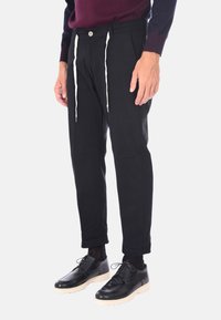 Pantaloni neri a sigaretta con chiusura a lacci e bottone. Presentano tasche anteriori e posteriori, abbinati a scarpe nere lucide.