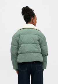 Casaco acolchoado verde em veludo cotelê com gola em algodão falso branco, apresentando um design acolchoado e textura canelada. Estilizado com jeans azul escuro.