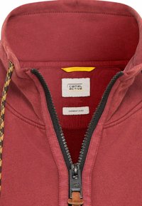 Polo rouge teint en pièce avec un col demi-zip, présentant un tissu texturé, une fermeture éclair contrastante et des étiquettes de marque à l'intérieur.