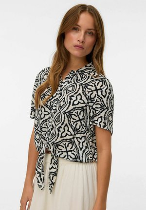 Chemise à manches courtes avec nouage à l'avant, présentant un motif géométrique noir et blanc sur un tissu léger. Col classique et coupe décontractée.