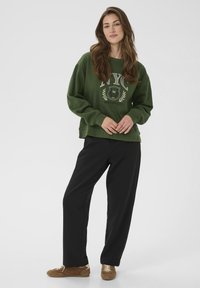 Felpa oversize verde in cotone con grafica e logo "NYC", abbinata a pantaloni neri a gamba larga e scarpe in suede marroni.