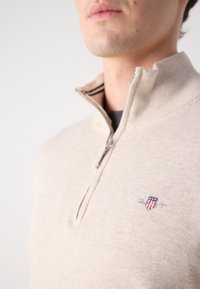 Suéter beige con cremallera y cuello acanalado, que presenta un pequeño logo bordado en el pecho. Textura suave, corte casual.