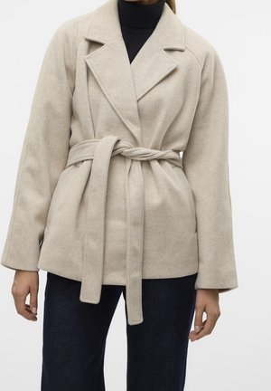 Femme portant un manteau en laine beige ceinturé sur un col roulé noir et un pantalon foncé, debout devant un fond blanc.