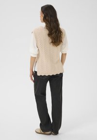 Gilet en tricot beige avec des motifs en losange, ourlet festonné, superposé sur une blouse blanche et porté avec un jean noir à jambes larges et des baskets beiges.