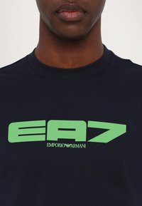 Jūrinės medvilnės marškinėliai su žaliu "EA7" ir "EMPORIO ARMANI" logotipu. Apvalus iškirpimas su trumpomis rankovėmis, glotni tekstūra.