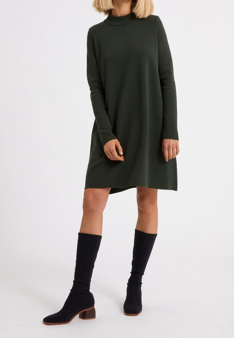 Femme portant une robe vert foncé à manches longues, longueur genou, avec des bottes noires à talons mi-mollet sur un fond clair uni.