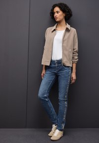 Chaqueta bomber beige con textura suave, camiseta blanca, jeans azules con detalles laterales y zapatillas beige. Diseño simple y corte ajustado.