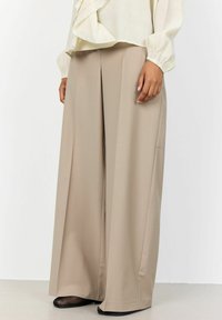 Beige weit geschnittene Hose mit glatter Textur und taillierter Passform. Mit einem gebügelten Knick versehen, ergänzt durch schwarze Mesh-Schuhe.