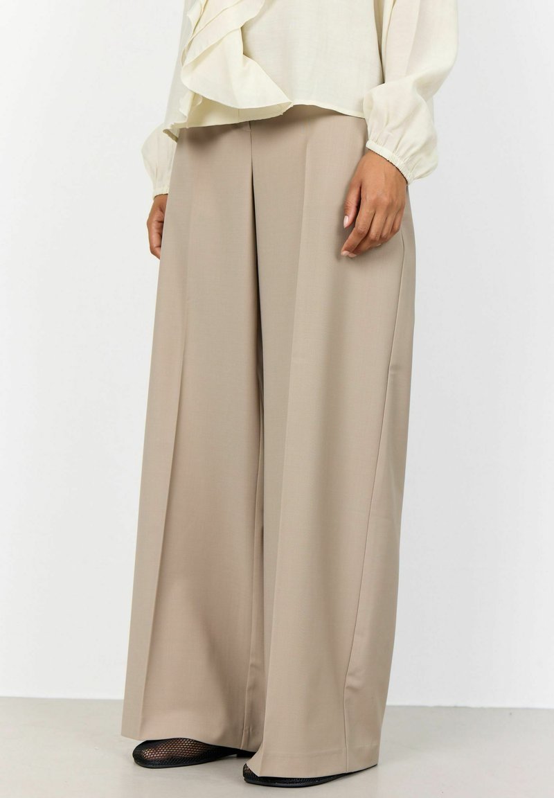 Beige weit geschnittene Hose mit glatter Textur und taillierter Passform. Mit einem gebügelten Knick versehen, ergänzt durch schwarze Mesh-Schuhe.