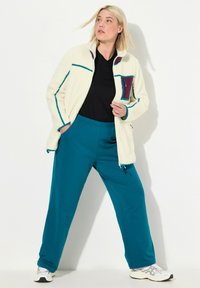 Kurtka polarowa w kolorze białym z niebieskimi przeszyciami, fioletowy detal kieszeni; zestawiona z niebieskimi spodami dresowymi i białymi butami sportowymi.