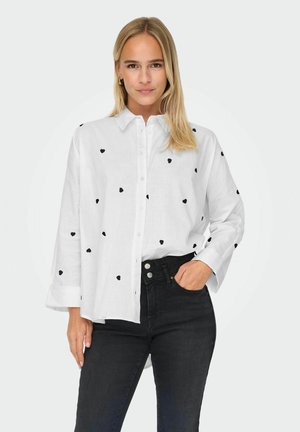 Camicia bianca con motivo a cuori neri, maniche lunghe e vestibilità rilassata, abbinata a jeans neri. Tessuto dalla texture liscia.