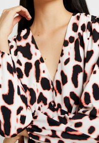 La blusa de estampado de leopardo cuenta con un profundo escote en V, mangas largas abullonadas y botones, destacando los colores negro, coral y crema. Textura suave.