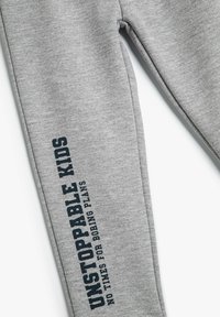 Pantalones de chándal grises fabricados con una tela suave que presentan un diseño de pierna recta. La pierna izquierda tiene un texto en azul oscuro: "NIÑOS INALCANZABLES SIN TIEMPO PARA PLANES ABURRIDOS."
