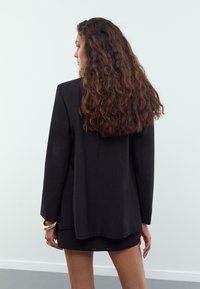 Eine Frau mit langen lockigen Haaren steht nach links gerichtet, trägt ein schwarzes Blazer und einen passenden Rock sowie goldene Armbänder und Ringe an ihrer linken Hand.