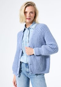 Femme portant un cardigan en tricot épais bleu clair par-dessus une chemise en denim à boutons et un jean bleu taille haute, se tenant devant un fond blanc.