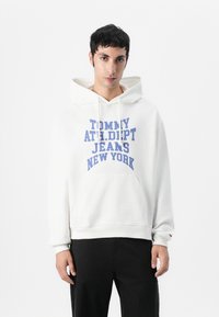 Sudadera blanca con gran texto azul que dice "TOMMY ATH. DEPT. JEANS NEW YORK." Presenta un bolsillo tipo canguro en la parte frontal y capucha con cordones.