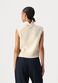 Strikket ærmeløs sweater i off-white med høj krave, ribbestrikket kant og tekstureret mønster. Vist fra ryggen, parret med mørke bukser.