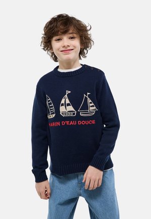 Garçon souriant aux cheveux bouclés portant un pull bleu marine avec des motifs de voiliers et du texte rouge, associé à un jean bleu clair et une chemise blanche en dessous.