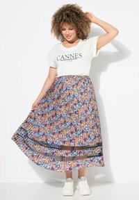 Gefalteter Rock mit floralem Muster in mehreren Farben auf dunklem Grund, kombiniert mit einem weißen T-Shirt mit dem Aufdruck "CANNES".