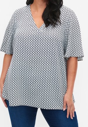 Frau trägt eine lockere schwarz-weiße V-Ausschnitt-Bluse mit flatternden Ärmeln und dunkelblaue Jeans, steht vor einem weißen Hintergrund.