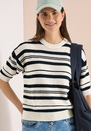 Junge Frau trägt ein schwarz-weiß gestreiftes kurzärmeliges Strickoberteil, eine hellblaue Baseballkappe, blaue Jeans und trägt eine marineblaue Tragetasche über einer Schulter.