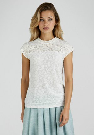 Junge Frau mit blonden Haaren, die ein weißes Spitzenoberteil mit kurzen Ärmeln und einen hellblauen, plissierten Rock vor einem schlichten Hintergrund trägt.