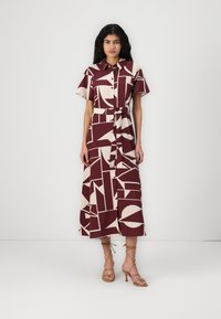 Robe à motifs marron et beige avec des manches courtes, fermeture pressionnée à l'avant, ceinture à la taille et longueur midi. Le mannequin porte des sandales dorées à lanières.