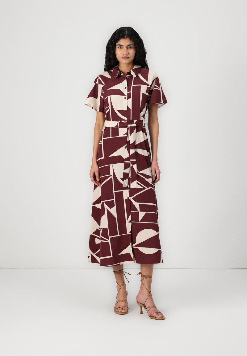Robe à motifs marron et beige avec des manches courtes, fermeture pressionnée à l'avant, ceinture à la taille et longueur midi. Le mannequin porte des sandales dorées à lanières.