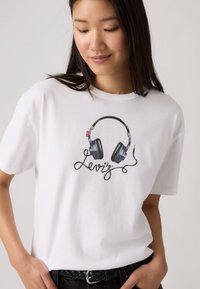 Γυναίκα με λευκή μπλούζα με εκτυπωμένα μαύρα ακουστικά και τη λέξη «Levi's» στο καλώδιο των ακουστικών, στέκεται με τα χέρια στις τσέπες.