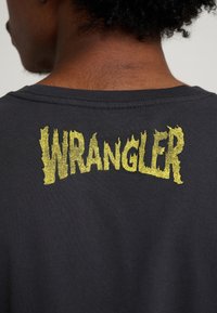 Mustasta puuvillaisesta t-paidasta löytyy keltainen, teksturoitu "WRANGLER"-logo selän pääntiellä. Pyöreä pääntie ja lyhyet hihat.