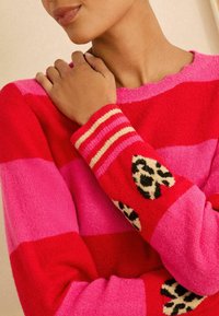 Kötött pulóver élénk vörös és rózsaszín csíkokkal, leopárd mintás akcentusokkal a hajtókán és csíkos mandzsettákon. Puha, texturált felület.