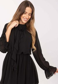 Czarna sukienka z szyfonu z długimi, falbaniastymi rękawami i wysokim dekoltem. Zdobiona kwiatowym akcentem przy szyi oraz rozkloszowanym dołem.
