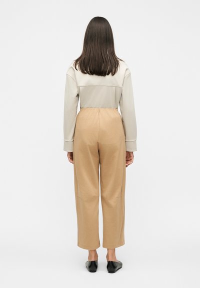 Γυναίκα που στέκεται με πλάτη, φοράει μπλούζα μπεζ μακρύ μανίκι, καφέ ψηλόμεσο παντελόνι με φαρδιά μπατζάκια και μαύρα επίπεδα παπούτσια σε λευκό φόντο.