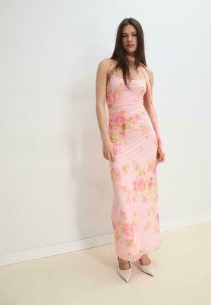 Frau steht vor einer weißen Wand und trägt ein langes ärmelloses Maxikleid mit rosa Blumenmuster und weiße spitze High Heels.