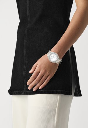 Montre en argent avec un cadran rond, bracelet transparent, portée sur une main. Haut en denim noir avec une fente sur le côté, associé à un bas côtelé blanc.