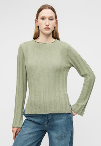 Νεαρή γυναίκα με ίσια ξανθά μαλλιά φοράει ριμπ πλεκτό, ανοιχτό πράσινο πουλόβερ με μακριά μανίκια και μπλε τζιν, ποζάροντας με το ένα χέρι στη μέση.