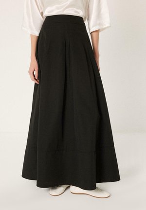 Γυναίκα φοράει μαύρη, μακριά φούστα Α γραμμή ψηλοκάβαλη, συνδυασμένη με λευκά παπούτσια και λευκό σατέν μπλουζάκι με μανίκια μέχρι τους αγκώνες.