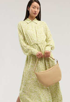 Žena v dlouhých rukávech, světle zelených vzorovaných šatech, držící béžovou kabelku s krátkým popruhem na jednotném pozadí.