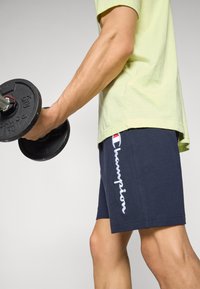 Marine treningsshorts med hvit "Champion" logo, kombinert med en lysegrønn t-skjorte. Personen holder en svart vekt i høyre hånd.
