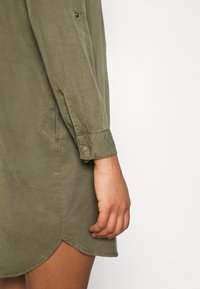 Gros plan sur le bras et la main d’une personne portant une robe-chemise à manches longues vert olive avec un revers de manche et un ourlet arrondi.