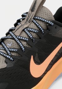 Close-up van een zwart en beige Nike hardloopschoen met gemusterde blauwe en zwarte veters en een oranje swoosh op een oranje zool.