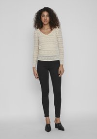 Blonde bluse i off-white med lange ærmer, V-hals og mønstret tekstur. Kombinert med svarte tettsittende bukser og spisse svarte sko.