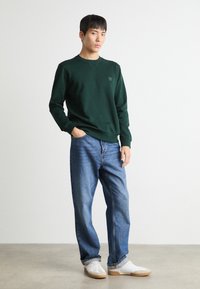 Donkergroen sweatshirt met een ronde halslijn, geribbelde manchetten en zoom; gecombineerd met lichtblauwe jeans en witte sneakers op een neutrale achtergrond.