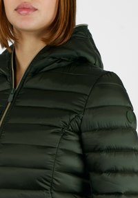 Giacca imbottita verde con cappuccio, caratterizzata da trapuntatura orizzontale, chiusura a zip e un distintivo rotondo sul braccio sinistro. Texture liscia.