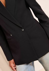 �Černý dvouřadý blazer s klopami, textilní struktura, dvě přední kapsy a černé knoflíky, spárovaný se světle modrými džíny.