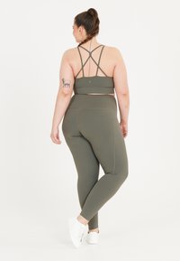 Oliivinvihreä urheiluliivi, jossa on ristikkäiset hihnat ja korkeat leggingsit. Sileä, joustava kangas, jossa on hienovaraisia ompelutietoja ja logoaksentteja.