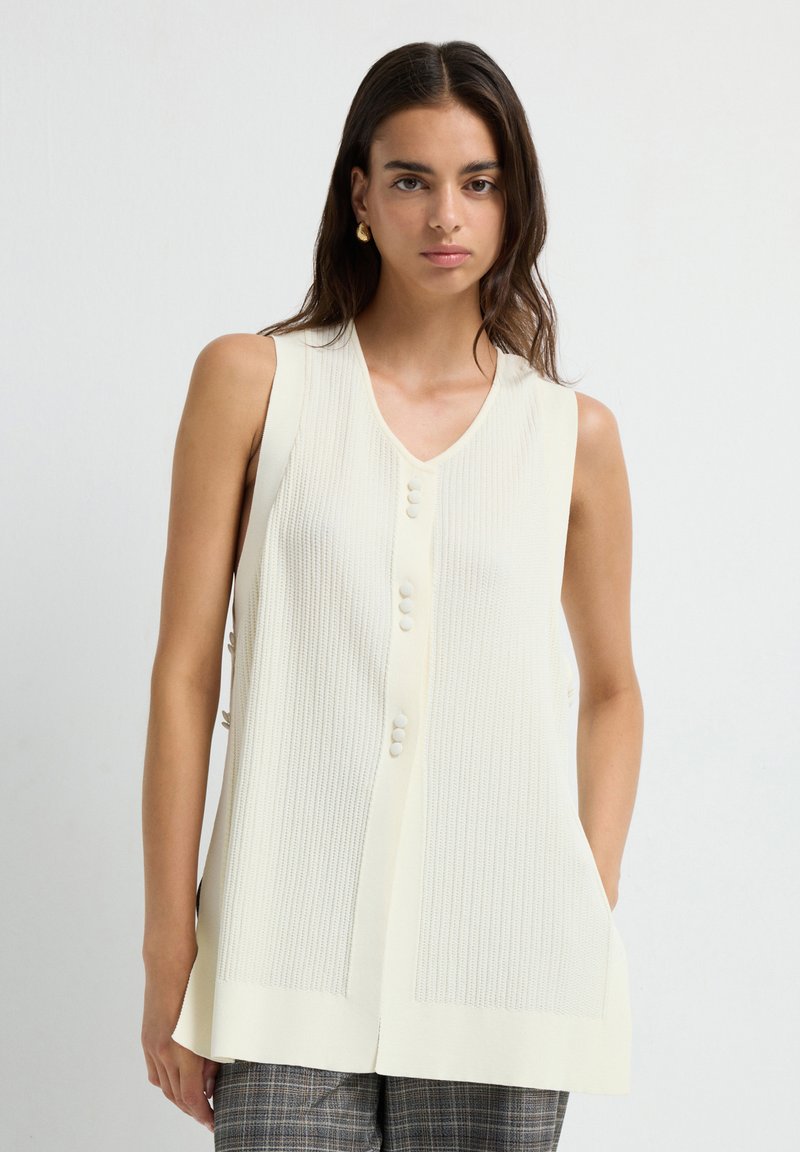 Lichtcreme mouwloze sweatervest met verticale ribbels, V-hals en een knoopsluiting aan de voorkant. Heeft een gestructureerde afwerking en zijsplitten.