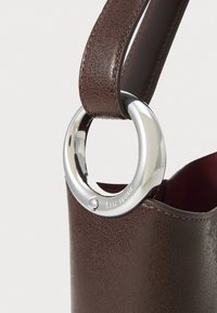 Brun læderhandtaske med tekstur og et sølvfarvet rundt hardwareaccent. Indersiden er foret med en ensfarvet burgunderfarve.