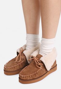 Chaussons en daim marron avec un col en fausse fourrure crème duveteux. Bout arrondi, surpiqûres décoratives et semelle en caoutchouc beige. Portés avec des chaussettes blanches à côtes.